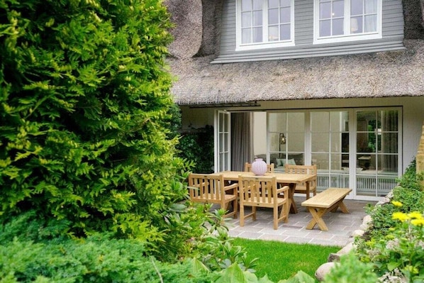 A house with a thatched roof, a patio with a table and chairs, and a garden with various plants. - Holiday home in Sylt with swimming pool (Sylt)