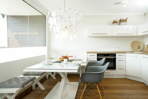 A modern kitchen with white cabinetry, a built-in oven, and a dining area with a glass table and grey chairs. - Holiday apartment with 1 bedroom (Westerland)
