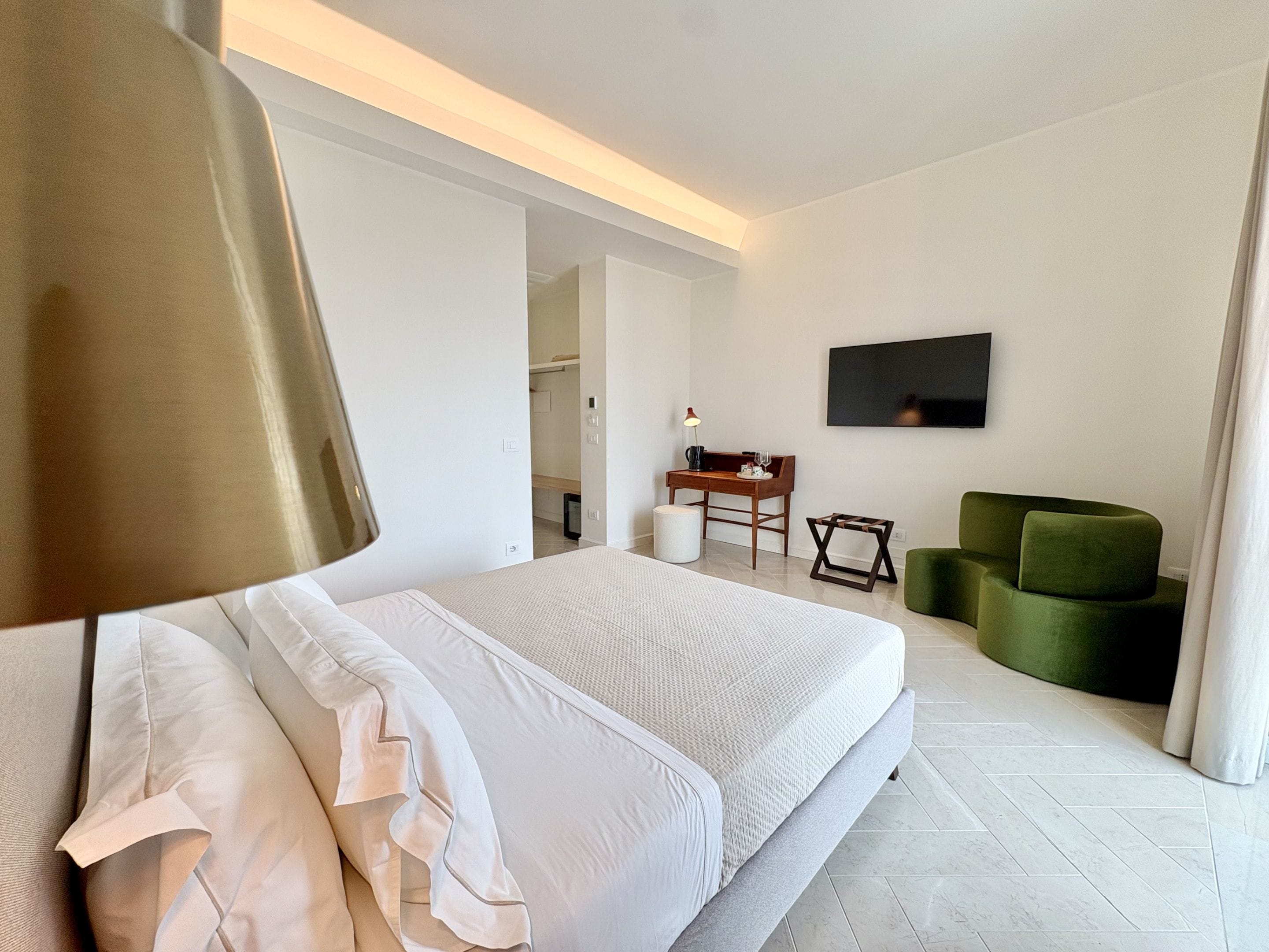 Quarto de hotel moderno com cama, escrivaninha, poltrona verde e TV de tela plana na parede.