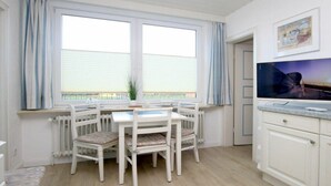 Een kamer met een eettafel en stoelen, een radiator, een raam met gordijnen en een televisie aan de muur.