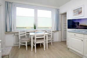 A room with a dining table and chairs, a radiator, a window with curtains, and a television on the wall. - Badestraße 90, Holiday Apartment 1 (Wyk auf Föhr)
