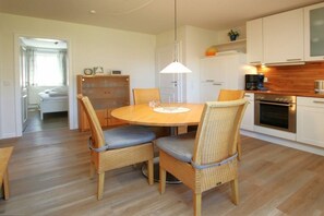 A dining area with a round table, wicker chairs with grey cushions, a pendant light, and a kitchen in the background. - Kiek Ut 9, Holiday Apartment 2 (Wyk auf Föhr)