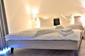 Ein ordentlich bezogenes Bett mit weißen Bettwäsche und Kissen, eine Nachttischlampe und ein dunkles Kopfteil.