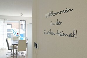 A room with a white wall featuring a welcome message in German. There is a dining area with a table and chairs visible through an open door. - Holiday home in Duhnen with Wi-Fi and parking (Duhnen)