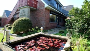 Een huis met een bakstenen gevel, een rood balkon, een klein vijvertje met waterlelies en een waterput.