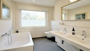 Een badkamer met een bad, twee wastafels, een toilet en een raam met zonwering.
