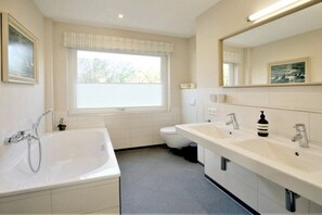 A bathroom with a bathtub, two sinks, a toilet, and a window with blinds. - Linge 4, Holiday Apartment 1, Hygge (Wrixum)