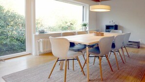 Een eetgedeelte met een houten tafel en stoelen, een groot raam met uitzicht op groen, en een hangende lamp.