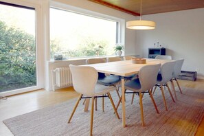 A dining area with a wooden table and chairs, a large window with a view of greenery, and a hanging light fixture. - Linge 4, Holiday Apartment 1, Hygge (Wrixum)