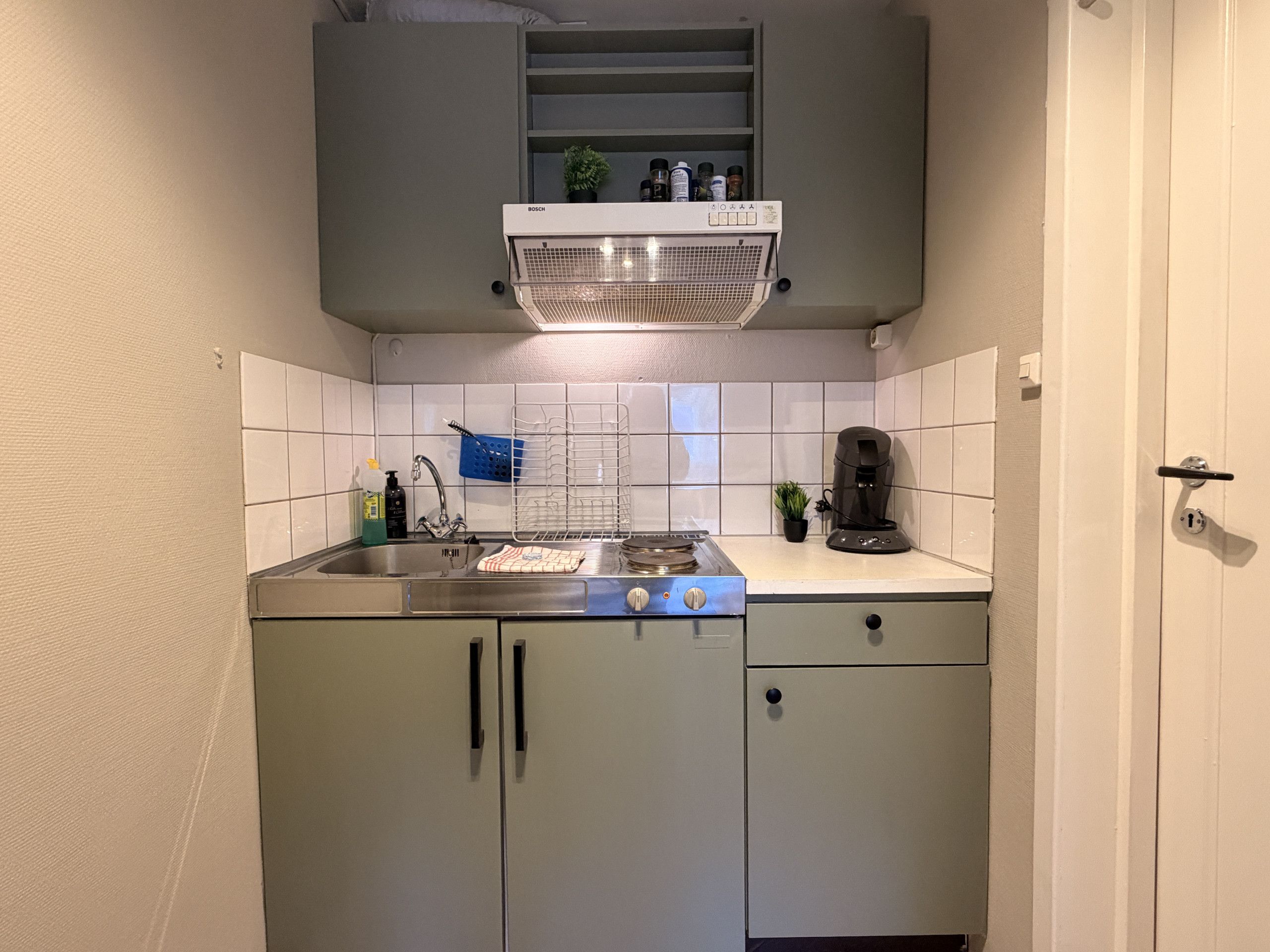 Una cocina compacta con estufa integrada, máquina de café y un pequeño estante sobre el fregadero.