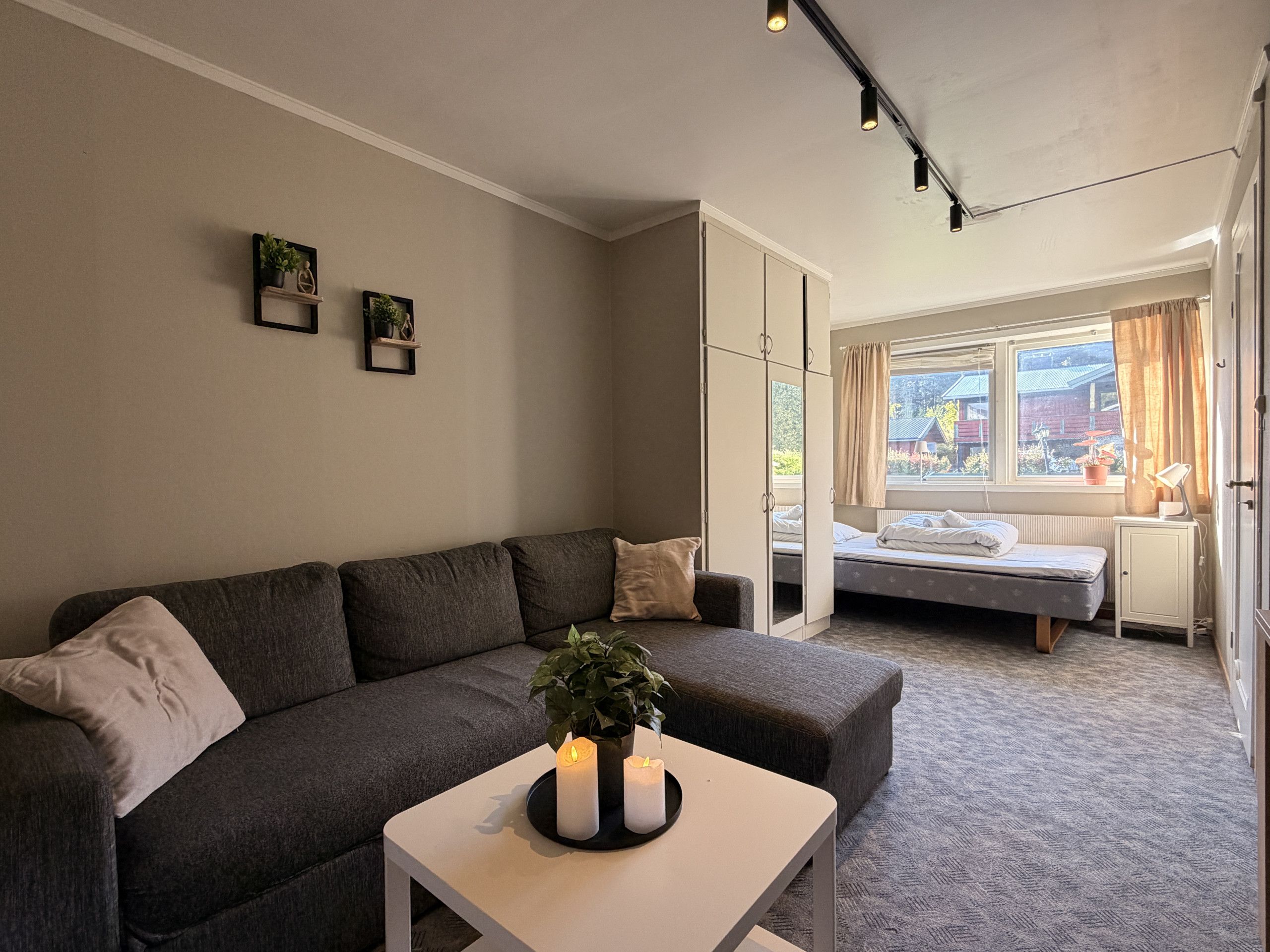 Un salon avec un canapé, une petite table avec des bougies, une fenêtre avec des rideaux et un lit à l’arrière-plan.
