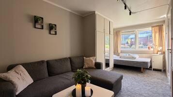 Uma sala de estar com sofá, uma mesinha com velas, janela com cortinas e uma cama ao fundo.
