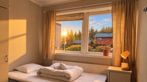 Quarto com cama, janela com cortinas, abajur e vista para um prédio vermelho lá fora.