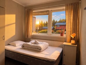 Ett sovrum med en säng, ett fönster med gardiner, en sänglampa och utsikt över en röd byggnad utanför.