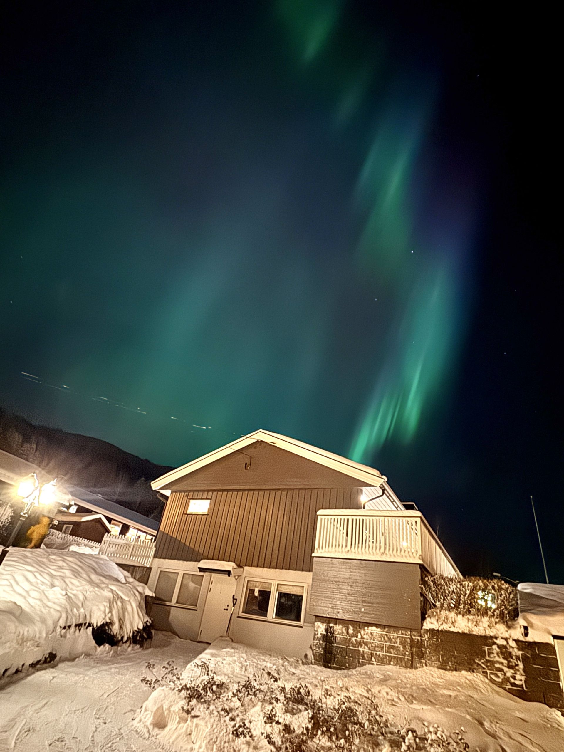 Um edifício com telhado coberto de neve e fachada de madeira, sob um céu noturno com aurora boreal visível.