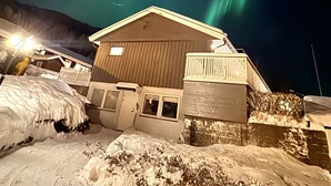 Um edifício com telhado coberto de neve e fachada de madeira, sob um céu noturno com aurora boreal visível.