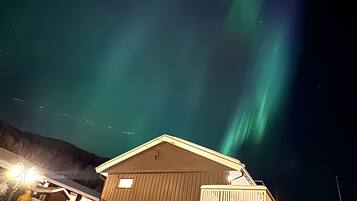 Um edifício com telhado coberto de neve e fachada de madeira, sob um céu noturno com aurora boreal visível.
