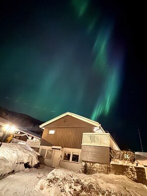 En byggnad med ett snötäckt tak och en träfasad, under en natthimmel där norrskenet syns.
