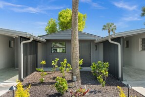 A modern two-story building with a central entrance, surrounded by a landscaped garden with young trees and shrubs. - Lake Patio + Firepit + Grill | Mins to Vanderbilt Beach | Fisherman's Cottage (Naples)
