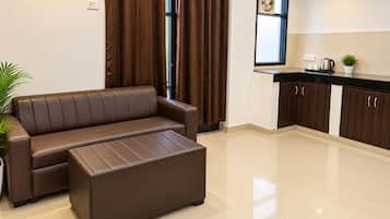 Ein modernes Wohnzimmer mit einem braunen Sofa und Hocker, ein Küchenbereich mit dunklen Holzschränken und ein Fenster mit Ventilator.