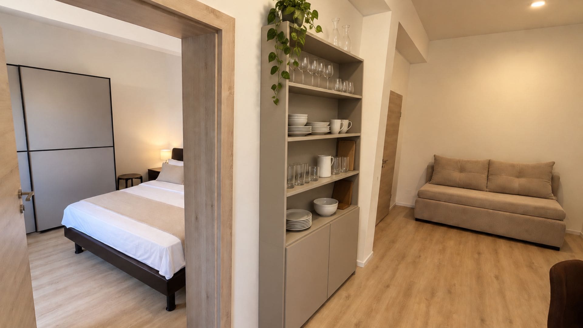 A modern bedroom with a bed, a sofa, and a built-in shelving unit with glassware and dishes.