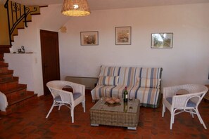 Una sala de estar con un sofá, dos sillones de mimbre, una mesa de centro con una botella y vasos, una escalera y tres cuadros en la pared.