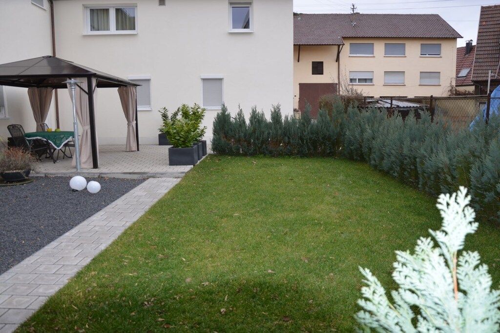 Een woonwijk met een gazon, een overdekt terras met stoelen en een rij altijdgroene bomen.