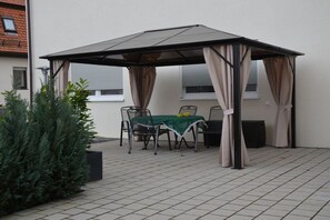 Ein überdachter Patio-Bereich mit Tisch und Stühlen, einer Topfpflanze und einem Gebäude im Hintergrund.