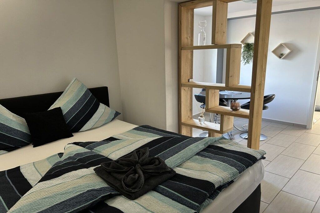 Una cama con ropa de cama a rayas y una toalla oscura, una escalera de madera y un comedor con mesa y sillas.