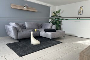 Uma sala de estar moderna com um sofá seccional cinza, uma mesa de centro de vidro, um tapete preto, um vaso branco e uma planta.