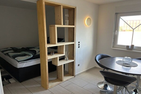 Quarto compacto com cama, estante de madeira, mesa de jantar com cadeiras e janela com vista.