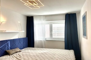 Quarto com cama, cabeceira, janela com cortinas e luminária fixada na parede.