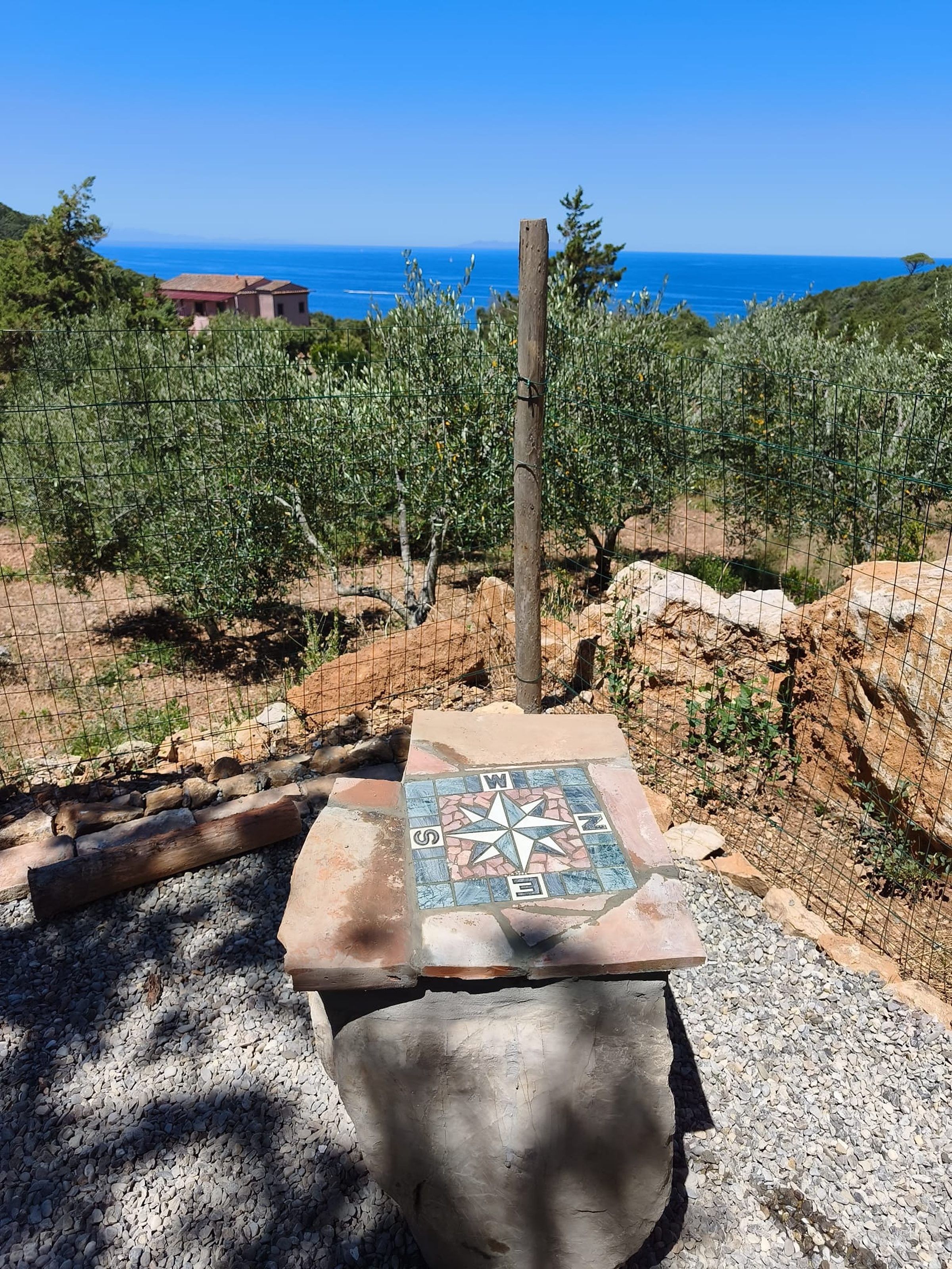 Un banc en pierre orné d’une mosaïque en forme de boussole, entouré d’une clôture en fil de fer, d’où l’on peut admirer la mer et, au loin, des oliviers.