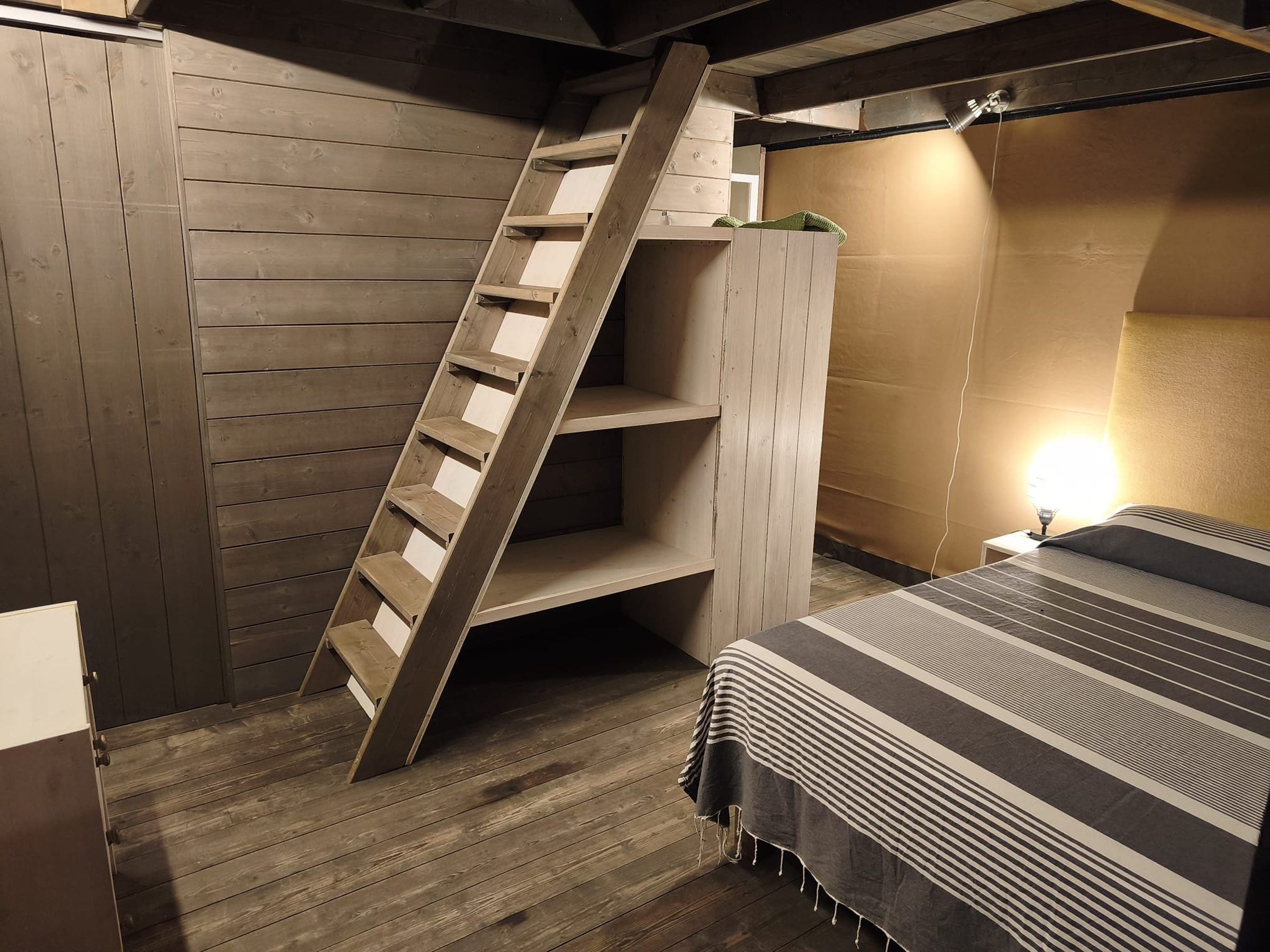 Une chambre avec une échelle en bois, un lit avec une literie à rayures et une table de chevet avec une lampe.