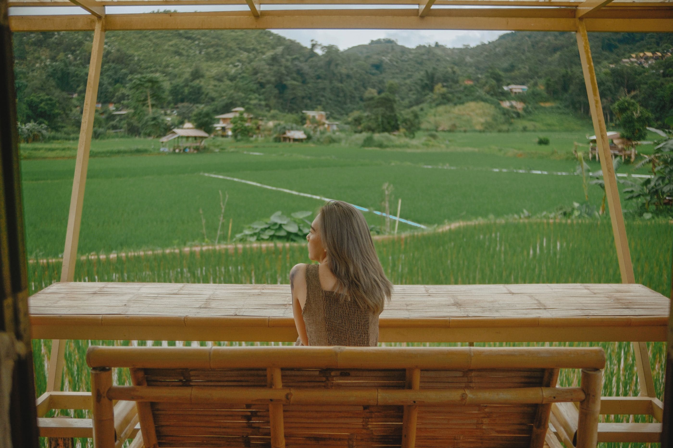 A person sitting on a wooden bench with a view of a green field and distant houses.