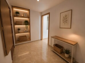 Un pasillo con una consola de madera, estantes con plantas y cestas, y una obra enmarcada en la pared.