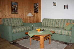 Ein Wohnzimmer mit einem grün gemusterten Sofa, einem Holztisch mit einer Obstschale und einer Lampe.