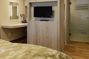 Quarto com cama, televisão montada num armário de madeira e espelho sobre a penteadeira.