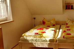 A bedroom with two beds, each with floral bedding and pillows. There is a window with a sheer curtain, and a bookshelf with books is visible in the background. - Holiday home on the Werra (Gerstungen\/OT Lauchröden)
