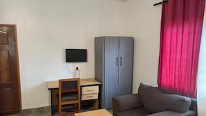 Una habitación con un escritorio de madera, una televisión, un armario gris, un sofá marrón y una cortina roja.