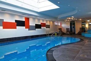 Piscina interior com design curvo, claraboia acima e área de estar com cadeiras.