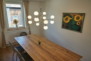 Un comedor con una mesa de madera, sillas, un candelabro, una ventana con cortinas y un cuadro de un girasol en la pared.