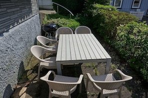 Un patio al aire libre con una mesa de madera y sillas de mimbre, rodeado de follaje y un muro de piedra.