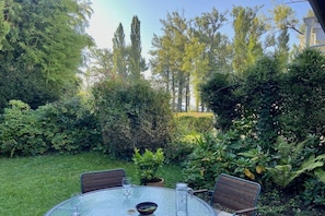 A round glass table with four chairs, surrounded by lush greenery and tall trees. - Rosenstrasse Gallery (Kressbronn)