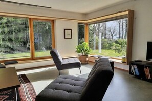 A modern living room with a sofa, a chair, a coffee table, and a television on a stand. There is a large window with blinds, a potted plant, and a bookshelf. - Rosenstrasse Gallery (Kressbronn)