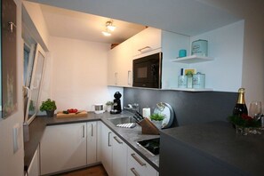 A modern kitchen with white cabinets, a black microwave, a sink, and a countertop with various items including a coffee maker, a cutting board with vegetables, and a bottle of champagne. - Alpenflair Holiday Apartment 106 (Oberstdorf)