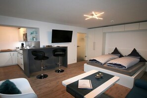 A modern living space with a kitchenette, a flat-screen TV, a sofa bed with patterned bedding, and a small table with a book. - Alpenflair Holiday Apartment 106 (Oberstdorf)