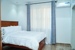 Un dormitorio con cama de madera, ropa de cama blanca, cortinas translúcidas y un equipo de aire acondicionado.