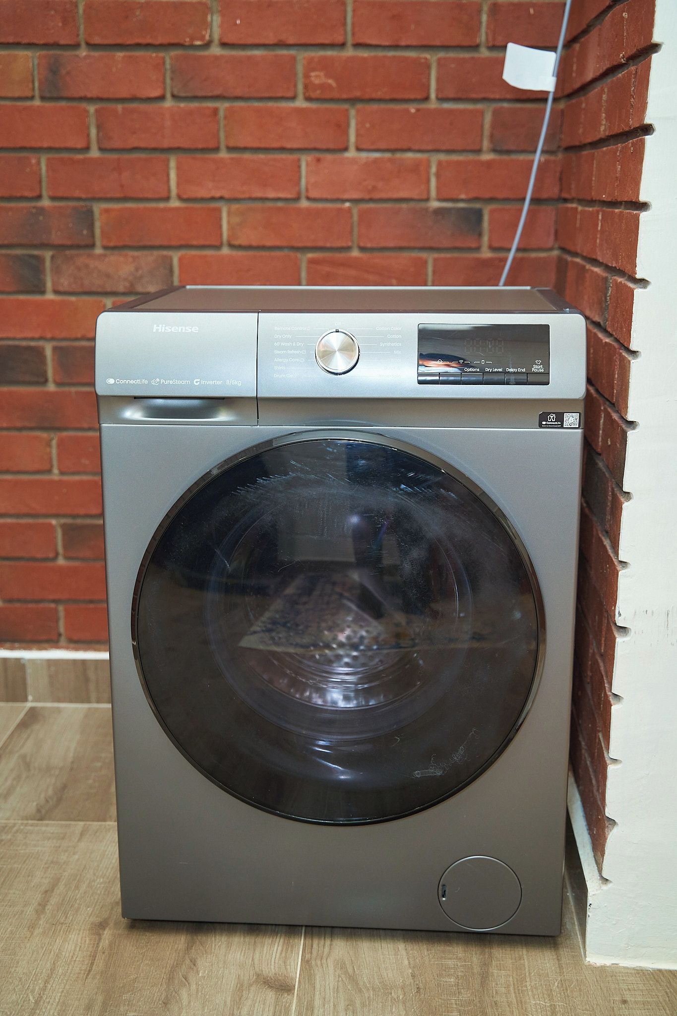 A front-loading Hisense washing machine with a control panel, set against a brick wall.