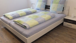 Een tweepersoonsbed met een lichtgekleurd hoofdeinde en voeteneinde, een nachtkastje met lade, en een stopcontact.